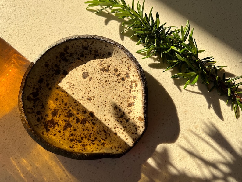 May include: A small, handmade ceramic dish with a speckled, off-white interior and a dark brown rim. The dish is partially filled with a golden-brown glaze. A sprig of fresh rosemary rests nearby, casting a shadow.