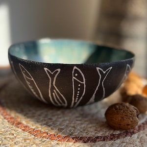 May include: A handmade black ceramic bowl with a teal interior and white fish designs around the exterior. The bowl is on a woven mat with walnuts and other nuts nearby.