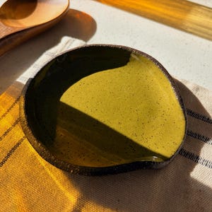 May include: An olive-green ceramic spoon rest with a dark brown rim. The spoon rest is on a yellow and white striped cloth, with a wooden spoon and a wooden spatula in the background. The spoon rest is approximately 13 cm in diameter.