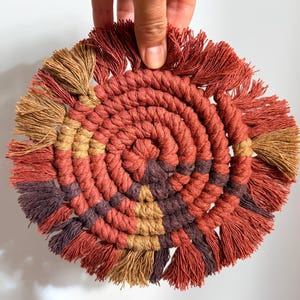 May include: A circular macramé coaster in shades of rust, brown, and tan. The coaster has a spiral design and a fringed edge. The handmade coaster is held by a hand against a white background.