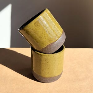 Op de afbeelding: Een set van drie keramische mokken met een gespikkelde, matte afwerking. De mokken zijn in crème, lichtgrijs en donkergrijs. Elke mok heeft een eenvoudig, minimalistisch ontwerp.