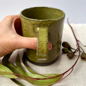 May include: A handmade ceramic mug with a speckled olive green glaze. The mug has a sturdy handle and a slightly textured surface. The mug is held by a hand, with eucalyptus leaves and seed pods in the background.