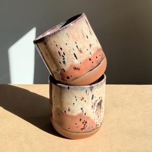 May include: Two handmade ceramic cups stacked on top of each other. The cups have a speckled beige and orange glaze with dark brown spots. The rims are a darker shade of brown. The cups are approximately 3.5 inches tall.