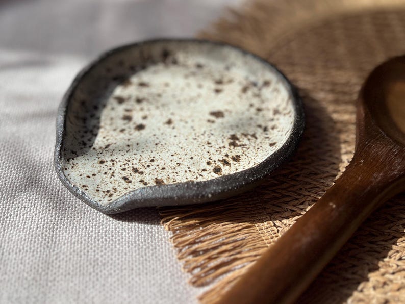 May include: A speckled ceramic spoon rest with a dark rim and a light-colored interior. The spoon rest is placed on a woven mat next to a wooden spoon. The speckled design adds a rustic touch to this kitchenware.