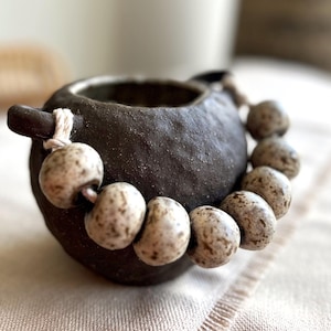 May include: A dark brown ceramic vessel with a small handle and a beaded necklace. The beads are speckled with light brown and white. The vessel is sitting on a light-coloured surface.