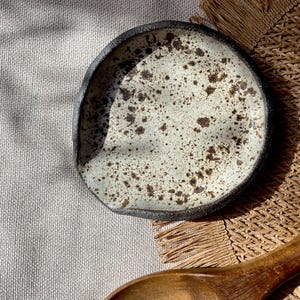 May include: A handmade ceramic spoon rest with a speckled off-white interior and a dark rim. The spoon rest is round with an organic shape. The background includes a wooden spoon and a woven mat.