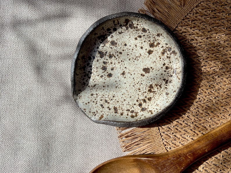 May include: A handmade ceramic spoon rest with a speckled off-white interior and a dark rim. The spoon rest is round with an organic shape. The background includes a wooden spoon and a woven mat.