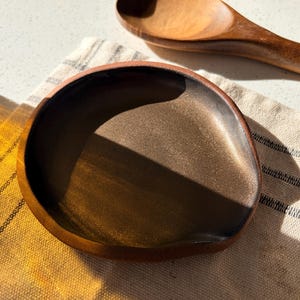 May include: A wooden spoon rest with a dark brown interior and a lighter brown rim. The spoon rest is on a striped beige and white cloth, with a wooden spoon resting nearby. The spoon rest is approximately 15 cm in diameter.
