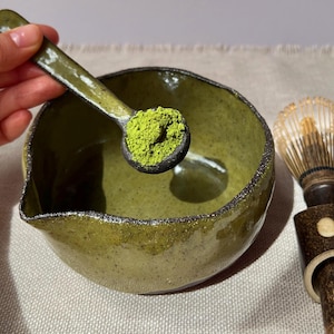 May include: A green ceramic matcha bowl with a spout, a matcha spoon holding green matcha powder, and a bamboo whisk. The bowl has a textured surface and a dark rim. The spoon and whisk have dark brown handles.