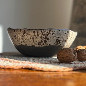 May include: A ceramic bowl with a speckled, off-white and brown exterior and a dark grey base. The bowl sits on a patterned mat with walnuts in front. The bowl's rim has a unique, uneven shape.