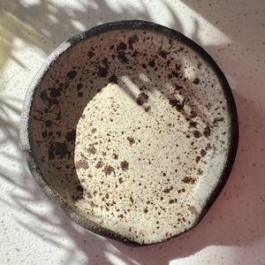 May include: A handmade ceramic spoon rest with a speckled cream-colored interior and a dark brown rim. The speckled pattern includes dark brown spots. The spoon rest is round with a slightly irregular shape.
