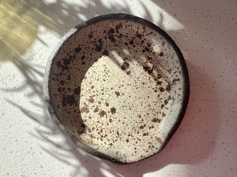 May include: A handmade ceramic spoon rest with a speckled cream-colored interior and a dark brown rim. The speckled pattern includes dark brown spots. The spoon rest is round with a slightly irregular shape.