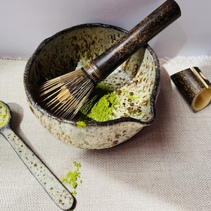 May include: A matcha tea set comprising a ceramic bowl with a bamboo whisk, a small ceramic spoon, and a bamboo tea holder. The bowl holds bright green matcha powder. The set is displayed on a neutral linen surface.