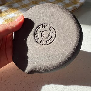 May include: A round, gray ceramic object with a stamped design in the center. The design features a leaf and the words "FIG & LIME" in a circular pattern. The object is held in a hand.