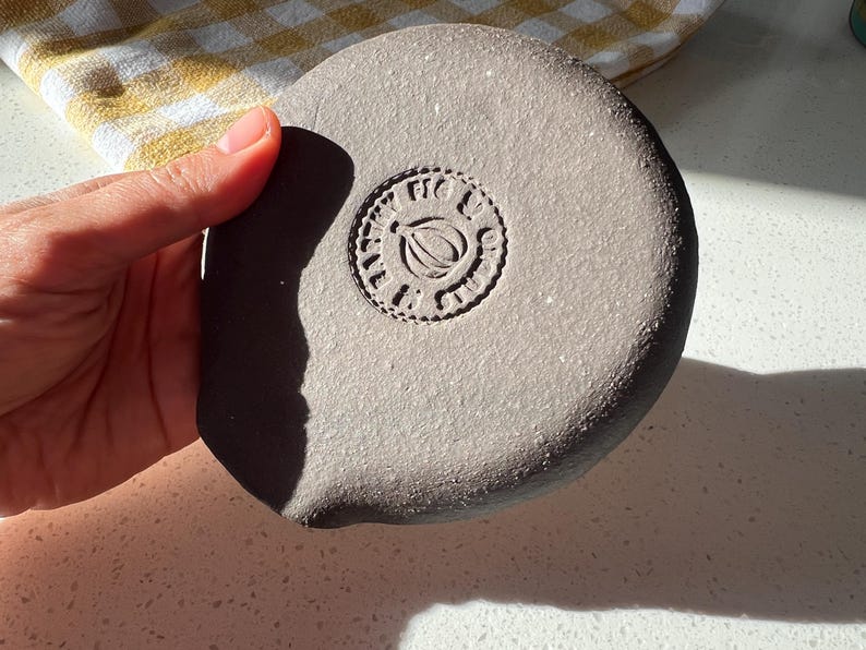 May include: A round, gray ceramic object with a stamped design in the center. The design features a leaf and the words "FIG & LIME" in a circular pattern. The object is held in a hand.