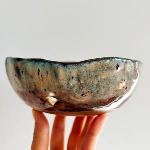 May include: A handcrafted ceramic bowl with an irregular, organic form. The bowl has a speckled glaze in brown, blue, and gold hues. The rim has a wavy edge, enhancing its artisanal charm. This decorative bowl is ideal for serving or display.