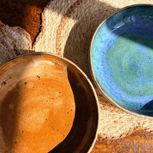 May include: Two handmade ceramic bowls. One bowl is a warm, earthy orange color, while the other is a vibrant blue and green. Both bowls have a rustic, textured appearance and are placed on a woven placemat.