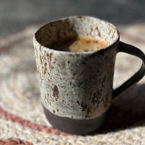 Puede incluir: Una taza de cerámica hecha a mano con un exterior moteado y texturizado en tonos beige y marrón. La base de la taza es de color marrón oscuro. La taza contiene un líquido oscuro, probablemente café o espresso. La taza está sobre una estera tejida.
