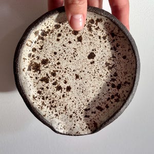 May include: A handmade ceramic spoon rest. The spoon rest has a speckled white interior with dark brown spots and a dark brown rim. The spoon rest is round with a small indentation on one side.
