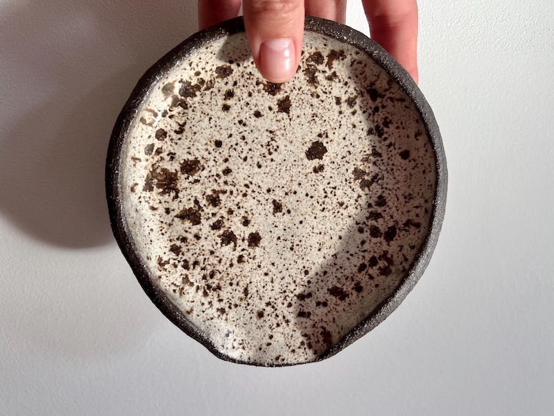 May include: A handmade ceramic spoon rest. The spoon rest has a speckled white interior with dark brown spots and a dark brown rim. The spoon rest is round with a small indentation on one side.
