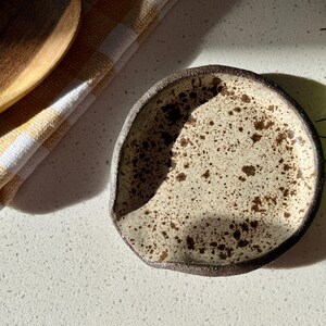 May include: A speckled ceramic spoon rest with a dark brown rim. The rest is off-white with brown speckles. A wooden spoon and a yellow and white checkered cloth are in the background. Rosemary sprigs are on the right.