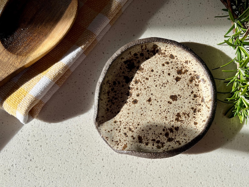 May include: A speckled ceramic spoon rest with a dark brown rim. The rest is off-white with brown speckles. A wooden spoon and a yellow and white checkered cloth are in the background. Rosemary sprigs are on the right.