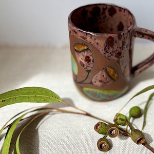 May include: A brown ceramic mug with a floral design. The mug has a handle and is decorated with stylized flowers in pink, yellow, and teal. A sprig of eucalyptus and seed pods are in the foreground. The mug is suitable for hot drinks.