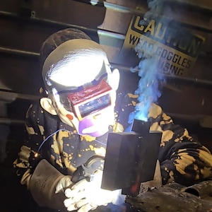 May include: A person wearing a clown mask and a black and tan bleach-splattered sweatshirt performs welding on a metal piece. A bright light emanates from the welding point, and a sign in the background reads CAUTION WEAR GOGGLES USING.
