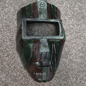 May include: A full-face mask featuring a textured, wood-like finish in alternating vertical stripes of dark brown and forest green. The mask includes a rectangular eye opening framed in silver, a small mouth slit, and a silver emblem at the top centre.