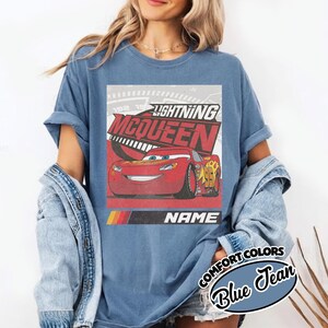 May include: A blue jean colored t-shirt featuring a graphic of the red race car Lightning McQueen. The design includes the text Lightning McQueen, a speedometer background, and a black bar with the word NAME above a yellow, orange, and red stripe.