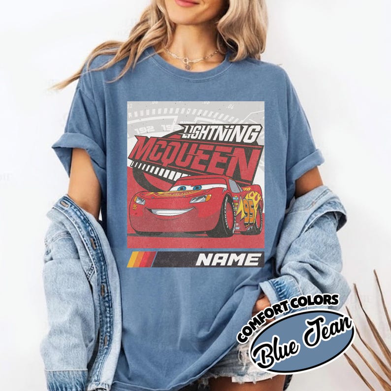 May include: A blue jean colored t-shirt featuring a graphic of the red race car Lightning McQueen. The design includes the text Lightning McQueen, a speedometer background, and a black bar with the word NAME above a yellow, orange, and red stripe.