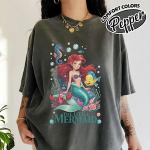 May include: Dark gray t-shirt featuring a colorful graphic of Ariel from The Little Mermaid, with Flounder, Sebastian, and a seahorse. The text "THE LITTLE MERMAID" is below the image. The shirt has the text "COMFORT COLORS Pepper" in a circular design.