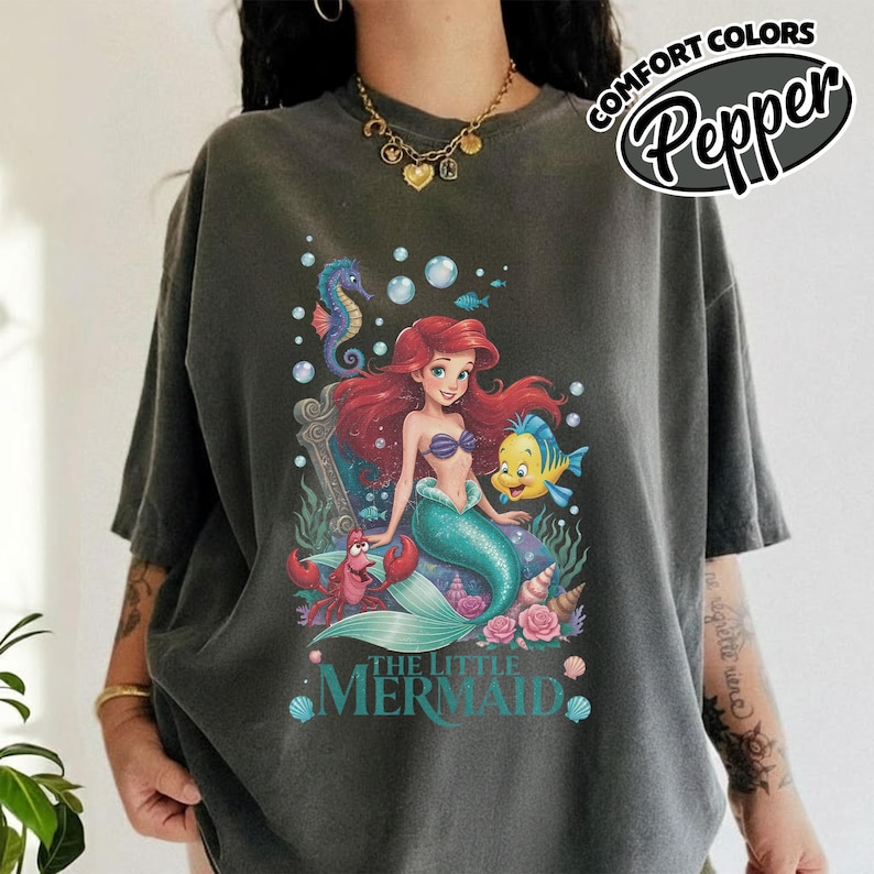 May include: Dark gray t-shirt featuring a colorful graphic of Ariel from The Little Mermaid, with Flounder, Sebastian, and a seahorse. The text "THE LITTLE MERMAID" is below the image. The shirt has the text "COMFORT COLORS Pepper" in a circular design.
