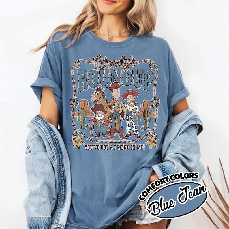 May include: Blue Comfort Colors t-shirt featuring a graphic of Woody's Roundup characters. The design includes the text "Woody's Roundup" and "You've Got a Friend in Me!" in a western-themed frame. The shirt is labeled "Blue Jean".