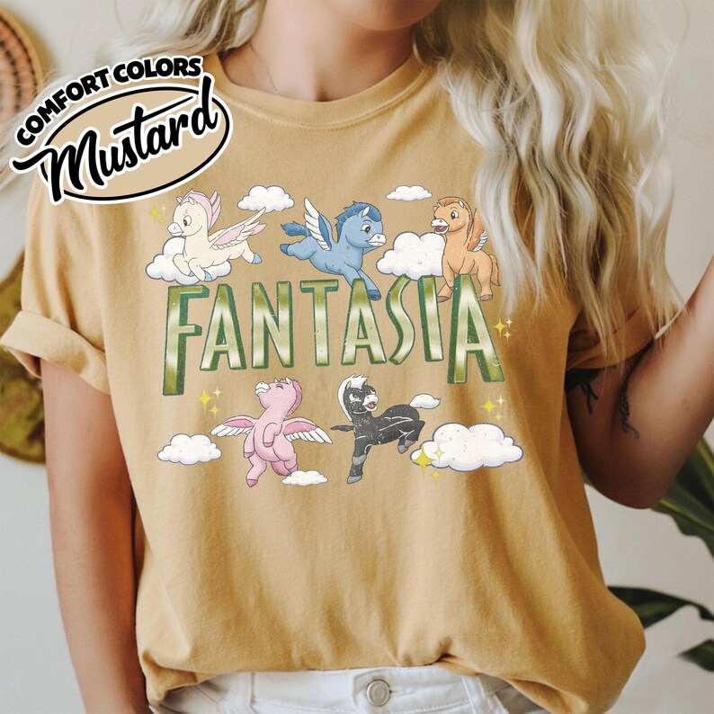 May include: Mustard-coloured t-shirt with the word "Fantasia" in green block letters, featuring cartoon pegasus characters and clouds. The shirt has rolled-up sleeves and a "Comfort Colors" logo.
