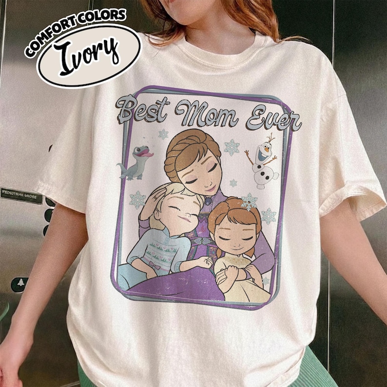 May include: Ivory-colored t-shirt with a graphic featuring the phrase "Best Mom Ever" and an illustration of a mother and two children. The design includes snowflakes and cartoon characters. The shirt has a Comfort Colors label.