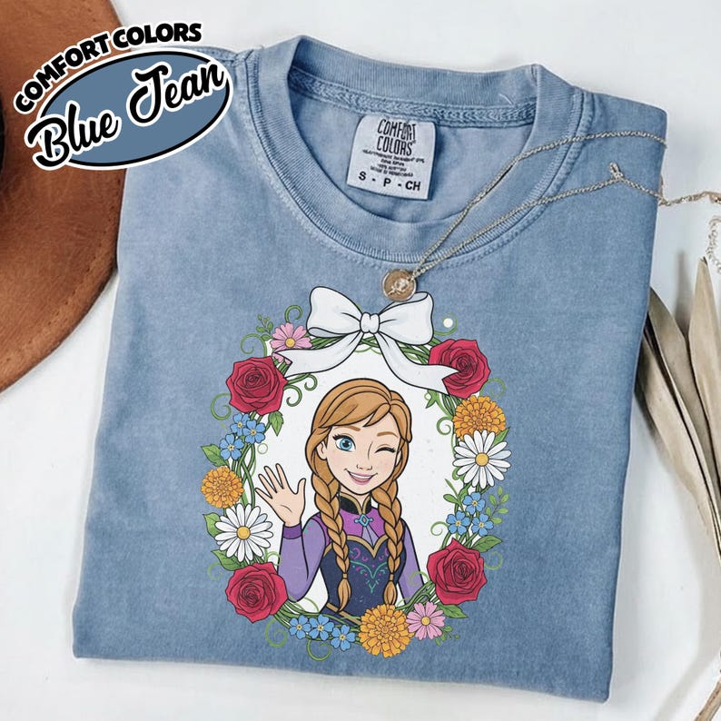Peut inclure: T-shirt bleu jean Comfort Colors avec un dessin anim&eacute; d'une femme faisant un clin d'&oelig;il et saluant, encadr&eacute; d'une couronne florale et d'un n&oelig;ud blanc. Le t-shirt a un col rond et des manches courtes. Un collier argent&eacute; avec un pendentif est drap&eacute; sur le t-shirt.