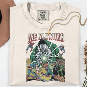 May include: Cream-colored t-shirt featuring a comic book-style graphic with the text "THE WAR WITHIN." The design includes illustrations of superheroes and villains in action, with a green-hooded figure at the center. A gold necklace with a star pendant is draped over the shirt.