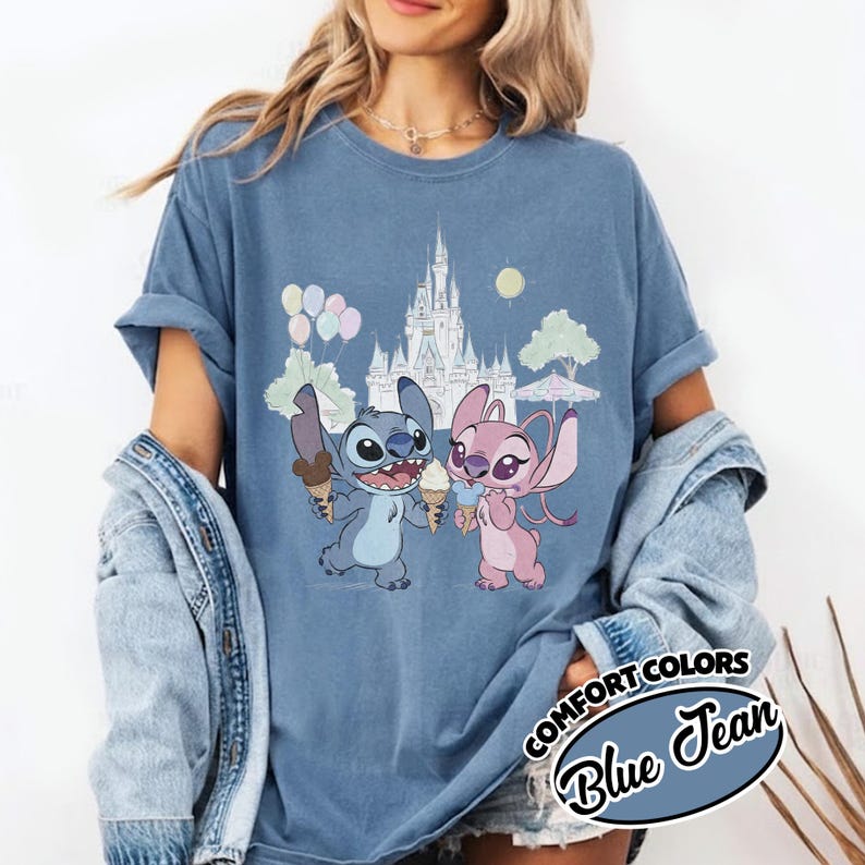 Puede incluir: Camiseta azul vaquero Comfort Colors con una ilustraci&oacute;n de dibujos animados de Stitch y Angel sosteniendo conos de helado frente a un castillo. El dise&ntilde;o incluye globos, &aacute;rboles y un sol. La camiseta tiene mangas enrolladas.