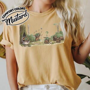 May include: Mustard-colored t-shirt featuring a cartoon illustration of Grogu in a forest setting. The graphic shows Grogu in various poses, with musical notes and a question mark. The text "COMFORT COLORS" and "Mustard" are printed on the top left.
