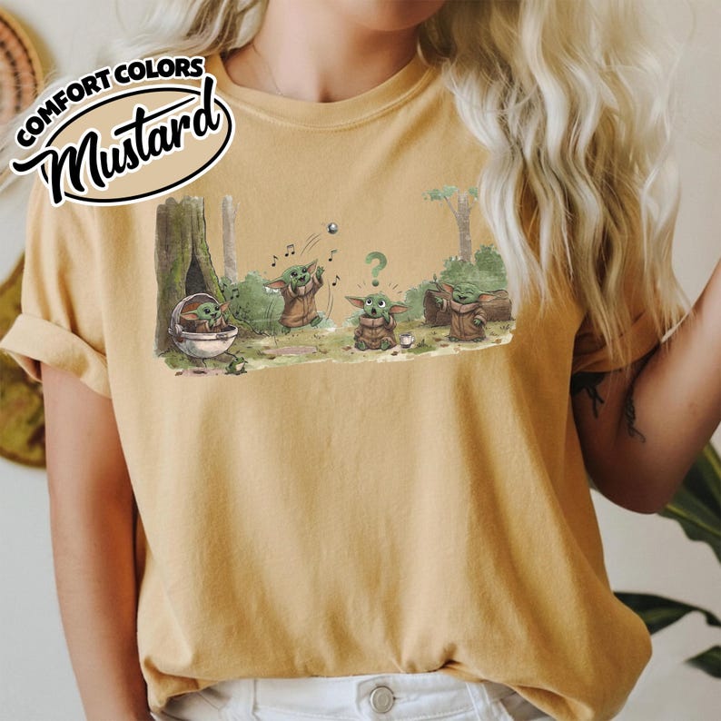 May include: Mustard-colored t-shirt featuring a cartoon illustration of Grogu in a forest setting. The graphic shows Grogu in various poses, with musical notes and a question mark. The text "COMFORT COLORS" and "Mustard" are printed on the top left.