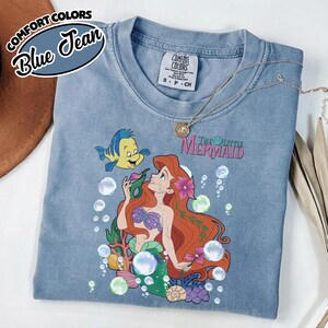 K&ouml;nnte beinhalten: Ein blaues T-Shirt in Jeansoptik mit einer Grafik von Arielle und Fabius aus Arielle, die Meerjungfrau, umgeben von Blasen, Korallen und Blumen. Das T-Shirt hat einen Rundhalsausschnitt und ein Comfort Colors Markenetikett am Innenkragen.
