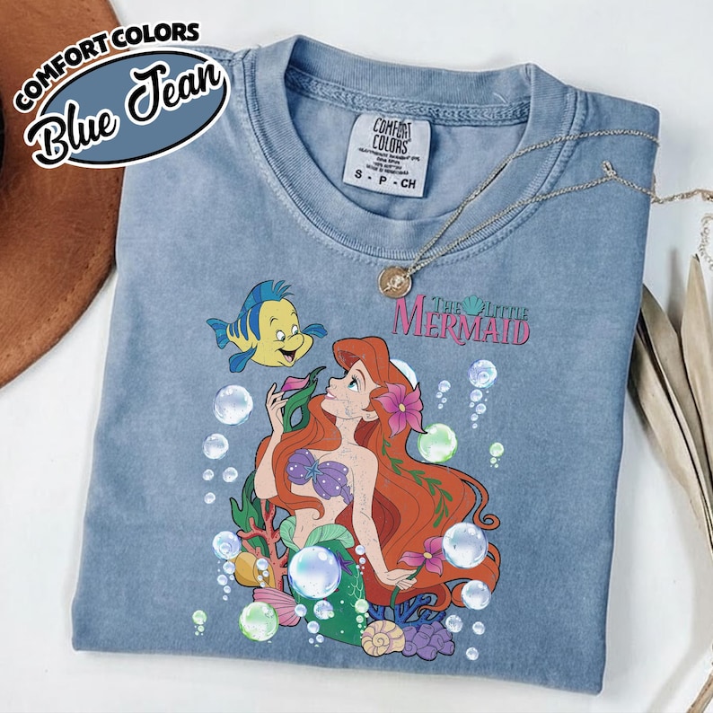K&ouml;nnte beinhalten: Ein blaues T-Shirt in Jeansoptik mit einer Grafik von Arielle und Fabius aus Arielle, die Meerjungfrau, umgeben von Blasen, Korallen und Blumen. Das T-Shirt hat einen Rundhalsausschnitt und ein Comfort Colors Markenetikett am Innenkragen.