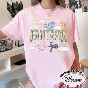 May include: Pale pink t-shirt with the word "FANTASIA" in green, and cartoon pegasus characters in different colours. The shirt also has the words "COMFORT COLORS" and "Blossom" in a circular design.