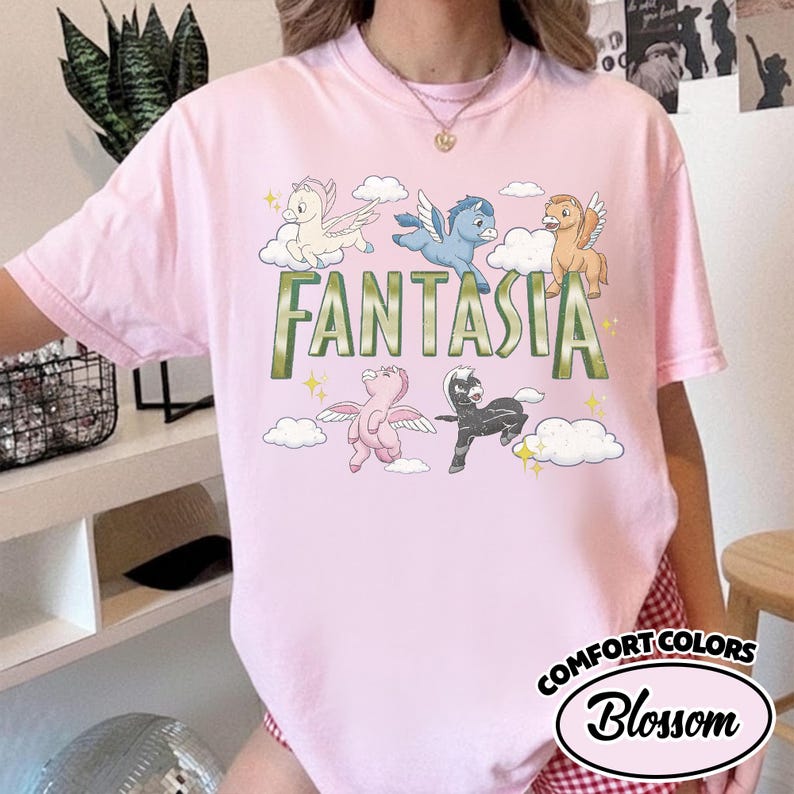 May include: Pale pink t-shirt with the word "FANTASIA" in green, and cartoon pegasus characters in different colours. The shirt also has the words "COMFORT COLORS" and "Blossom" in a circular design.