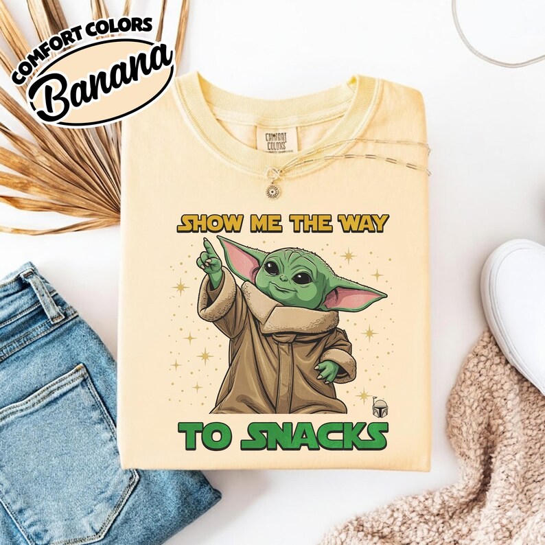 May include: A light yellow sweatshirt with the text "SHOW ME THE WAY TO SNACKS" and a graphic of a green alien character. The sweatshirt has a crew neck and long sleeves. The brand name "Comfort Colors" is printed on the top left.