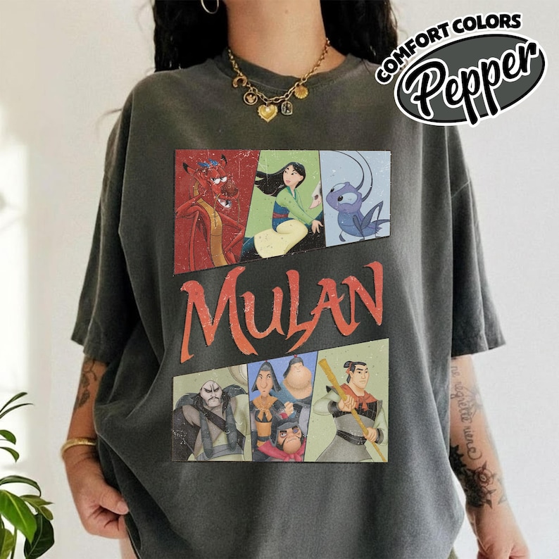 Puede incluir: Camiseta gris oscuro con un gr&aacute;fico de personajes de la pel&iacute;cula animada Mulan. El dise&ntilde;o incluye im&aacute;genes de Mushu, Mulan y otros personajes, con la palabra "Mulan" en naranja. La camiseta tiene mangas cortas y un corte holgado.