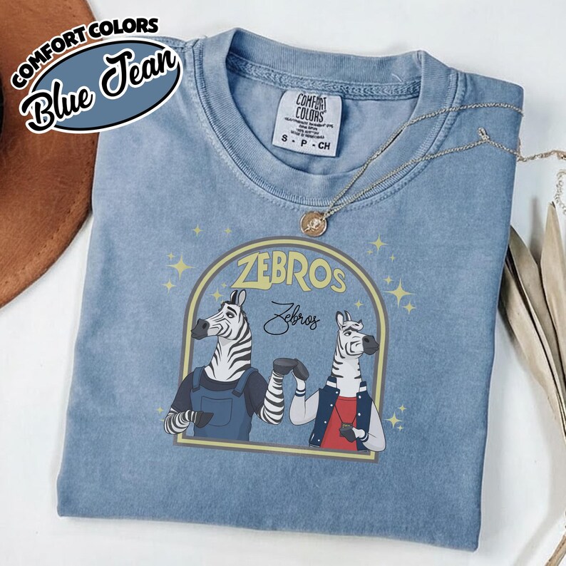 May include: Blue Comfort Colors t-shirt with a graphic of two zebras in a yellow archway. The text "ZEBROS" and "Zebros" is above the zebras. The shirt has a crew neck and short sleeves.
