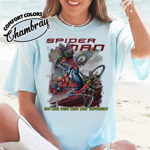 Op de afbeelding: Lichtblauw T-shirt met een Spider-Man afbeelding. Het ontwerp toont Spider-Man die vecht tegen de Green Goblin, met de woorden "SPIDER-MAN" en "WITH GREAT POWER COMES GREAT RESPONSIBILITY". Het shirt heeft de tekst "COMFORT COLORS Chambray" linksboven.