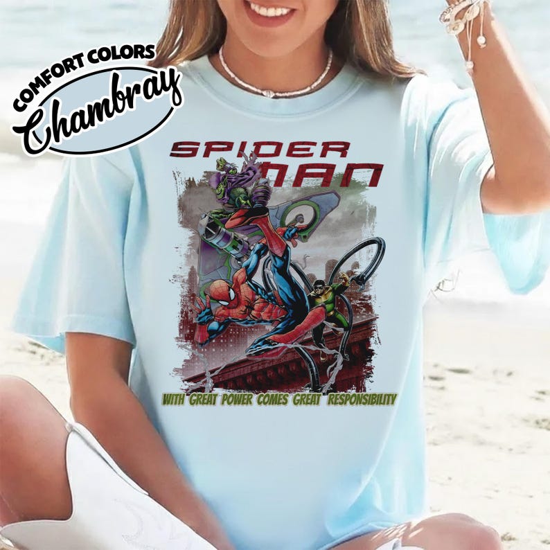 Op de afbeelding: Lichtblauw T-shirt met een Spider-Man afbeelding. Het ontwerp toont Spider-Man die vecht tegen de Green Goblin, met de woorden "SPIDER-MAN" en "WITH GREAT POWER COMES GREAT RESPONSIBILITY". Het shirt heeft de tekst "COMFORT COLORS Chambray" linksboven.